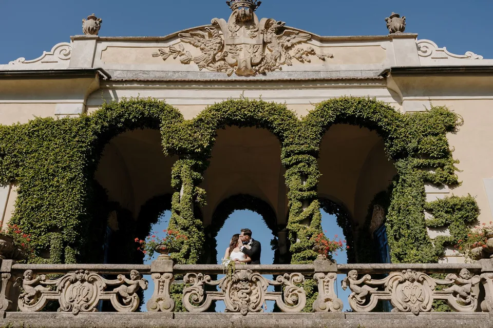 Wide view of Lyndal and Lachlan framed by Villa del Balbianello’s ivy-covered arches and carved stone balustrade beneath a blue sky, at Lake Como in Lombardy, Italy, for an Elopement Collective elopement. Photograph by George Bowden.