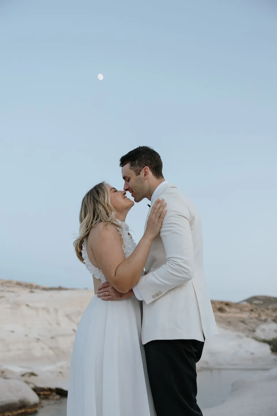 Georgina and Brad kissing in profile beneath the moon, with pale rock formations behind them, at Sarakiniko on Milos in the Cyclades, Greece, during an Elopement Collective elopement. Photograph by George Bowden.