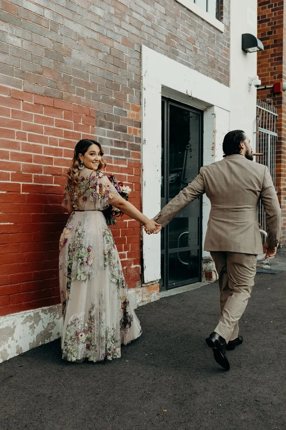 Daniel stepping away while holding Victoria’s hand beside a red brick building, in Fortitude Valley, Brisbane, for an Elopement Collective elopement. Photo by Tessa Shannon.