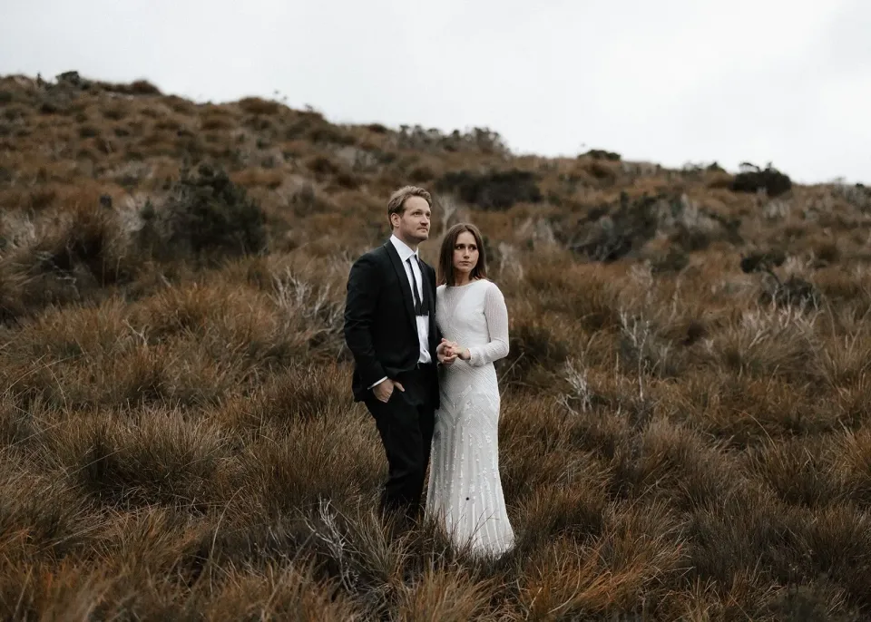 April and Josh standing among golden tussock grasses beneath the hills, in Cradle Mountain, Tasmania, for an Elopement Collective elopement. Photo by Bulb Creative.