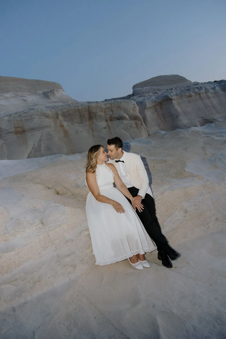 Georgina leaning close to Brad for a kiss as they sit together on pale rocks, at Sarakiniko on Milos in the Cyclades, Greece, during an Elopement Collective elopement. Photograph by George Bowden.