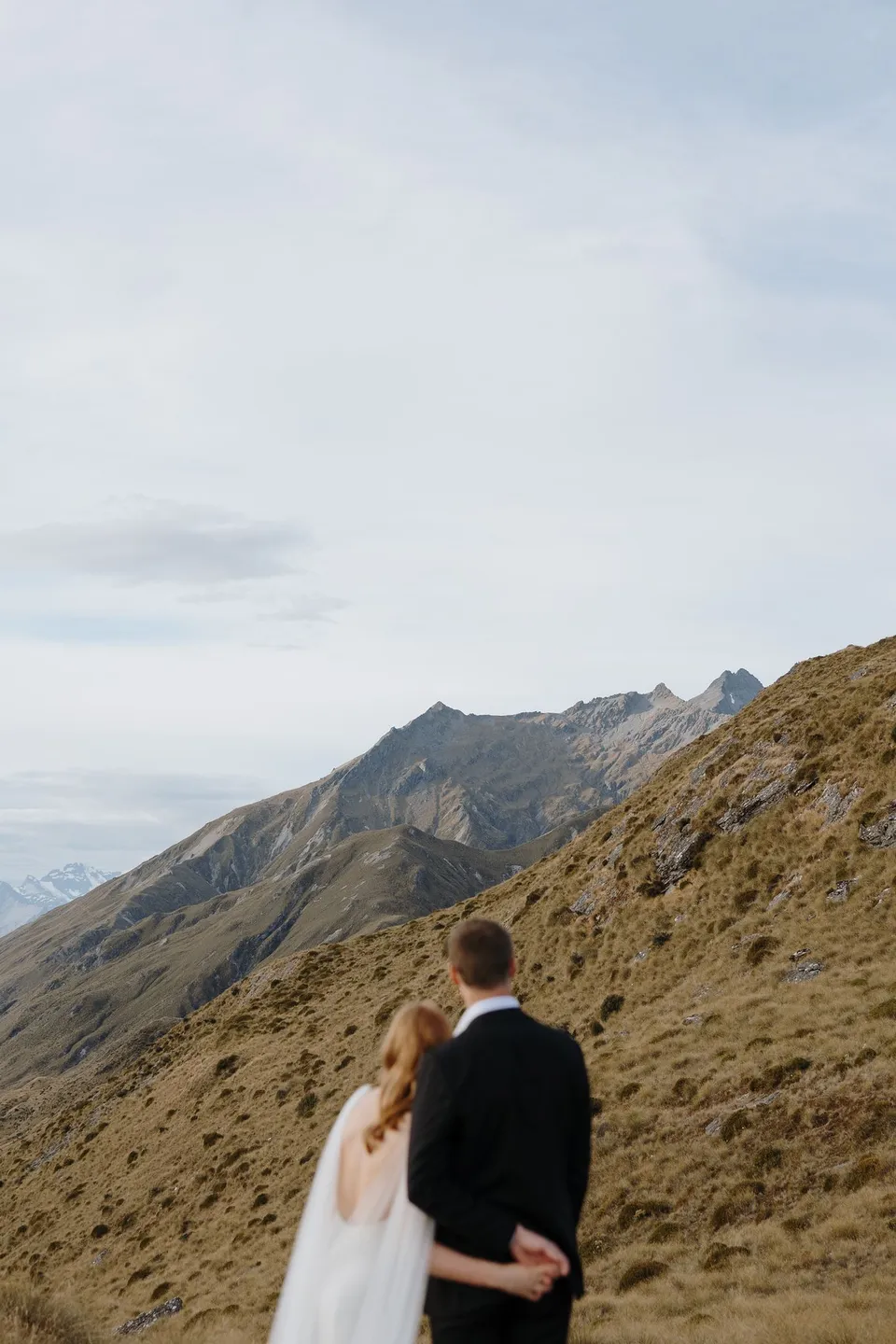 Neve and Hamish standing arm in arm beneath a broad sky, looking towards the mountain ridges, on Big Geordie above Queenstown, New Zealand, for an Elopement Collective Queenstown elopement. Photo by George Bowden.