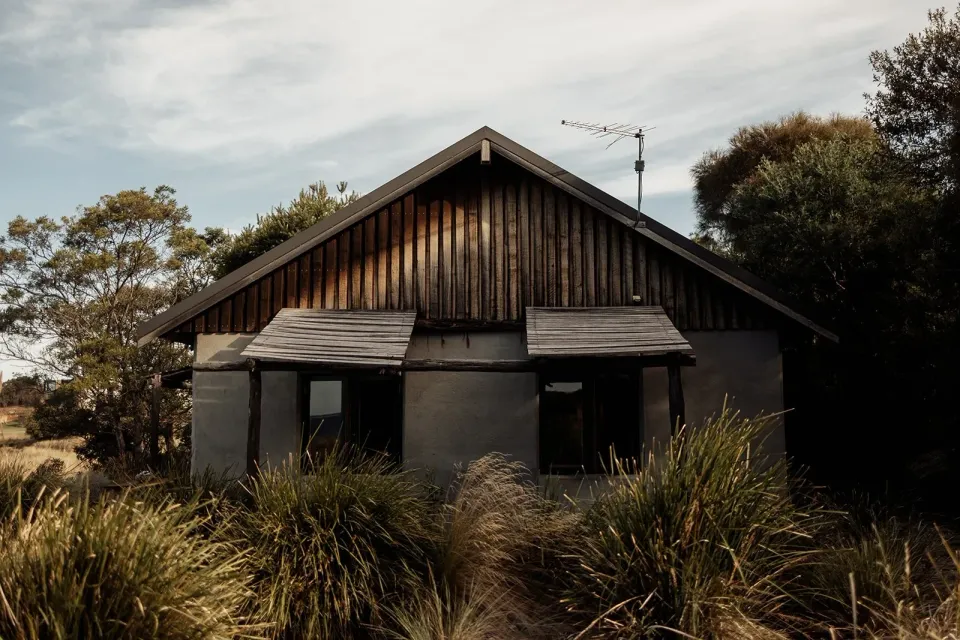 A timber cabin with shuttered windows nestled among coastal shrubs and grasses.