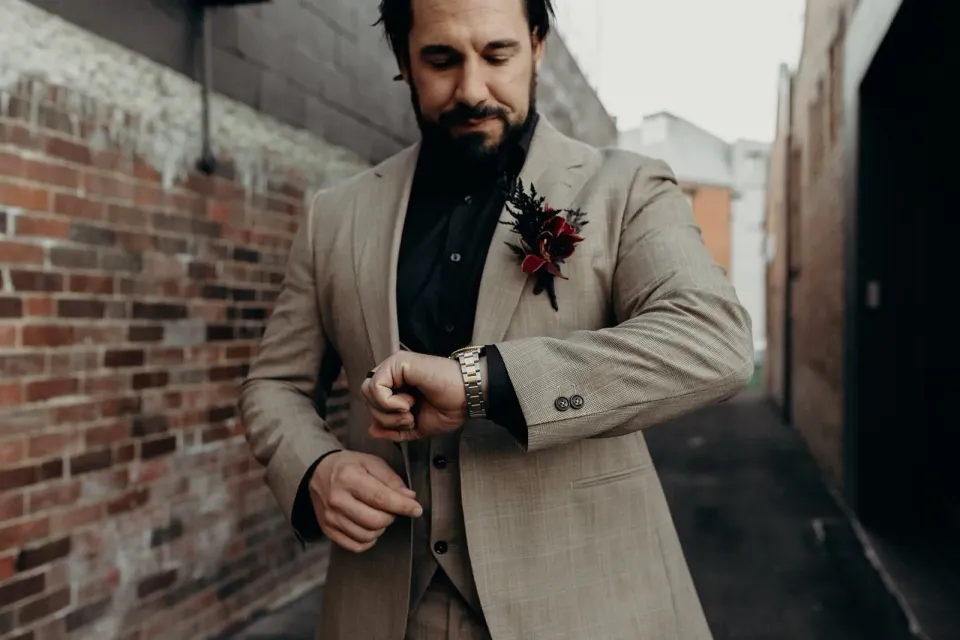 Daniel adjusting his wristwatch, with his beige suit and burgundy floral buttonhole visible, in Fortitude Valley, Brisbane, for an Elopement Collective elopement. Photo by Tessa Shannon.