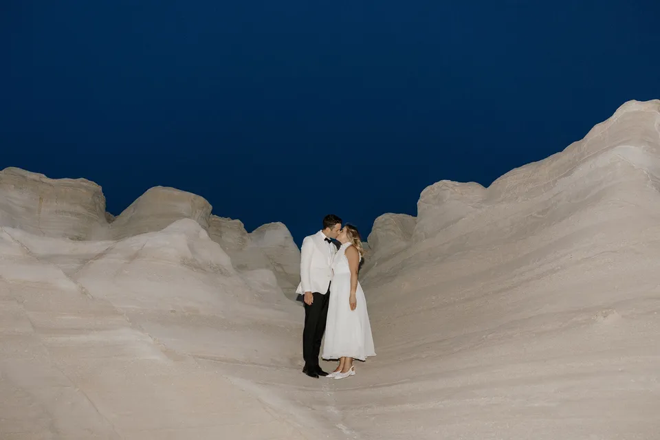 Georgina and Brad kissing and embracing in a curved white rock gully beneath a deep blue twilight sky, at Sarakiniko on Milos in the Cyclades, Greece, during an Elopement Collective elopement. Photograph by George Bowden.