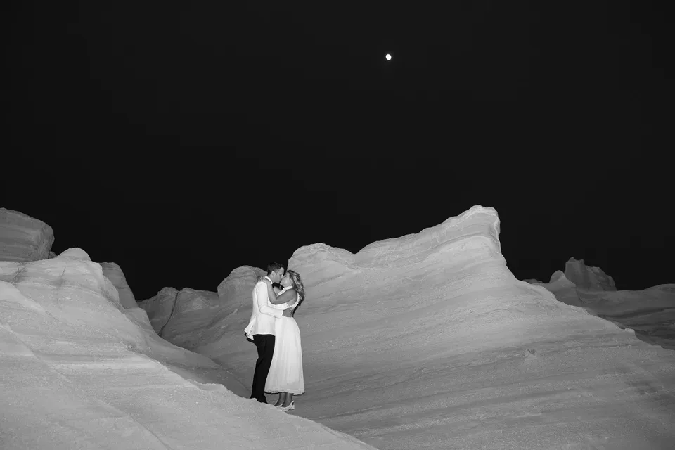 Black and white photograph of Georgina and Brad embracing in a white rock gully beneath a dark sky and visible moon, at Sarakiniko on Milos in the Cyclades, Greece, during an Elopement Collective elopement. Photograph by George Bowden.