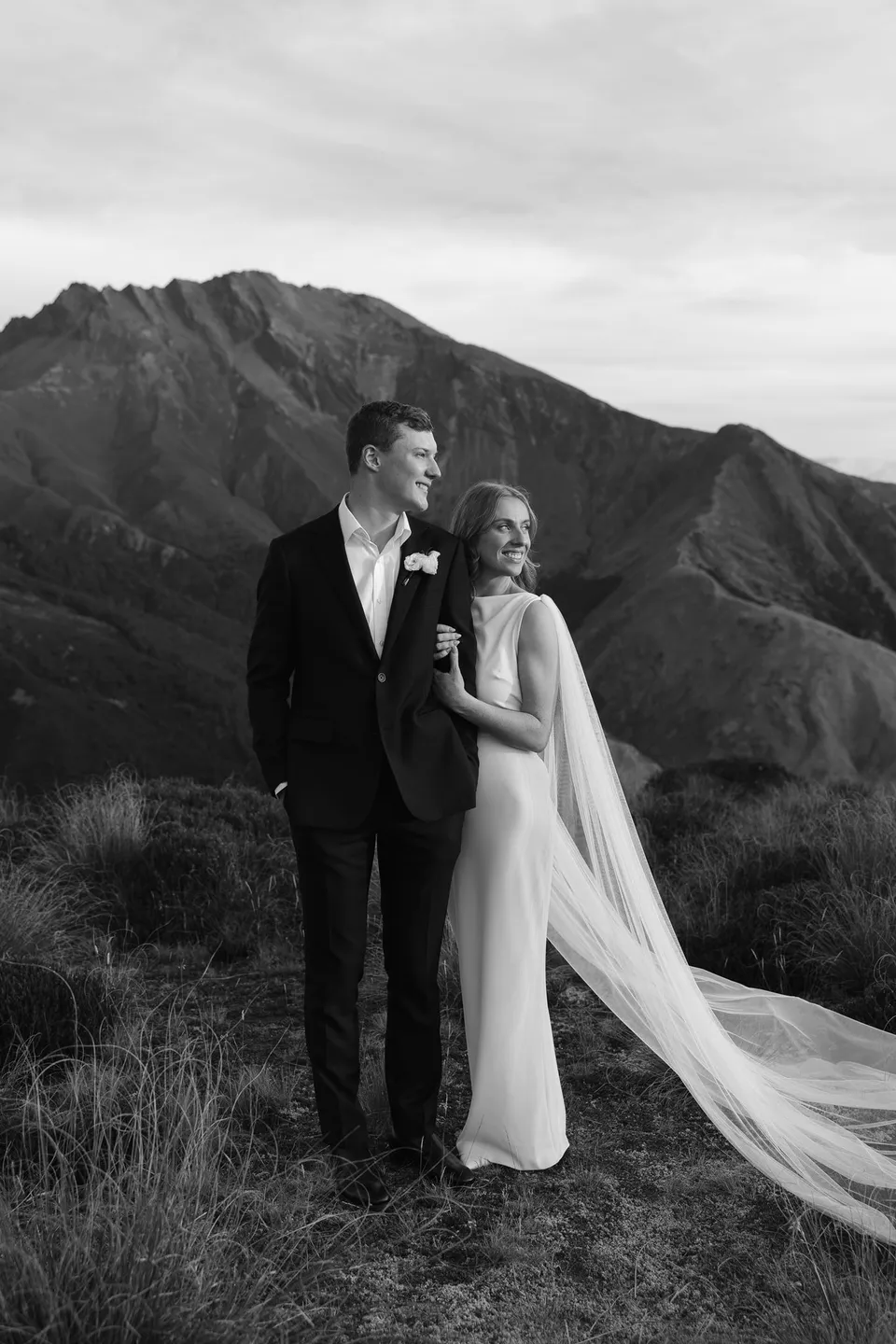 A black and white portrait of Neve leaning against Hamish, with her sheer cape draped across the tussock and mountains behind them, on Big Geordie above Queenstown, New Zealand, for an Elopement Collective Queenstown elopement. Photo by George Bowden.