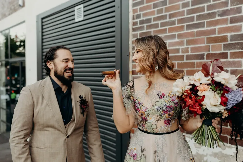 Victoria laughing with a cigar in hand beside Daniel and her bouquet by Mrs Gibbons Flowers, in Fortitude Valley, Brisbane, for an Elopement Collective elopement. Photo by Tessa Shannon.
