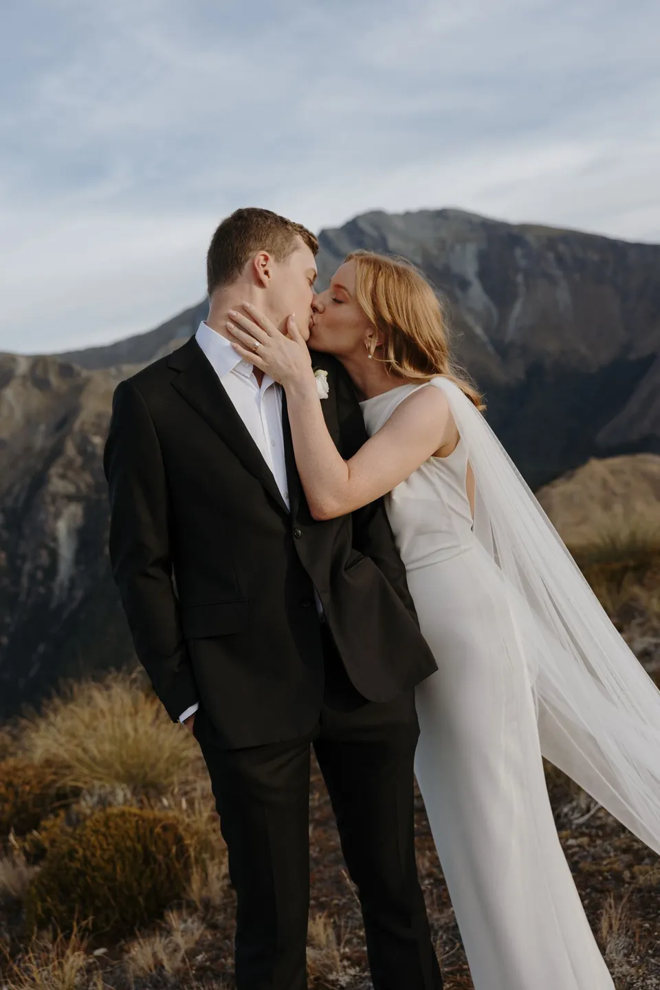 Neve resting a hand on Hamish’s neck as they kiss against a mountain backdrop, on Big Geordie above Queenstown, New Zealand, for an Elopement Collective Queenstown elopement. Photo by George Bowden.