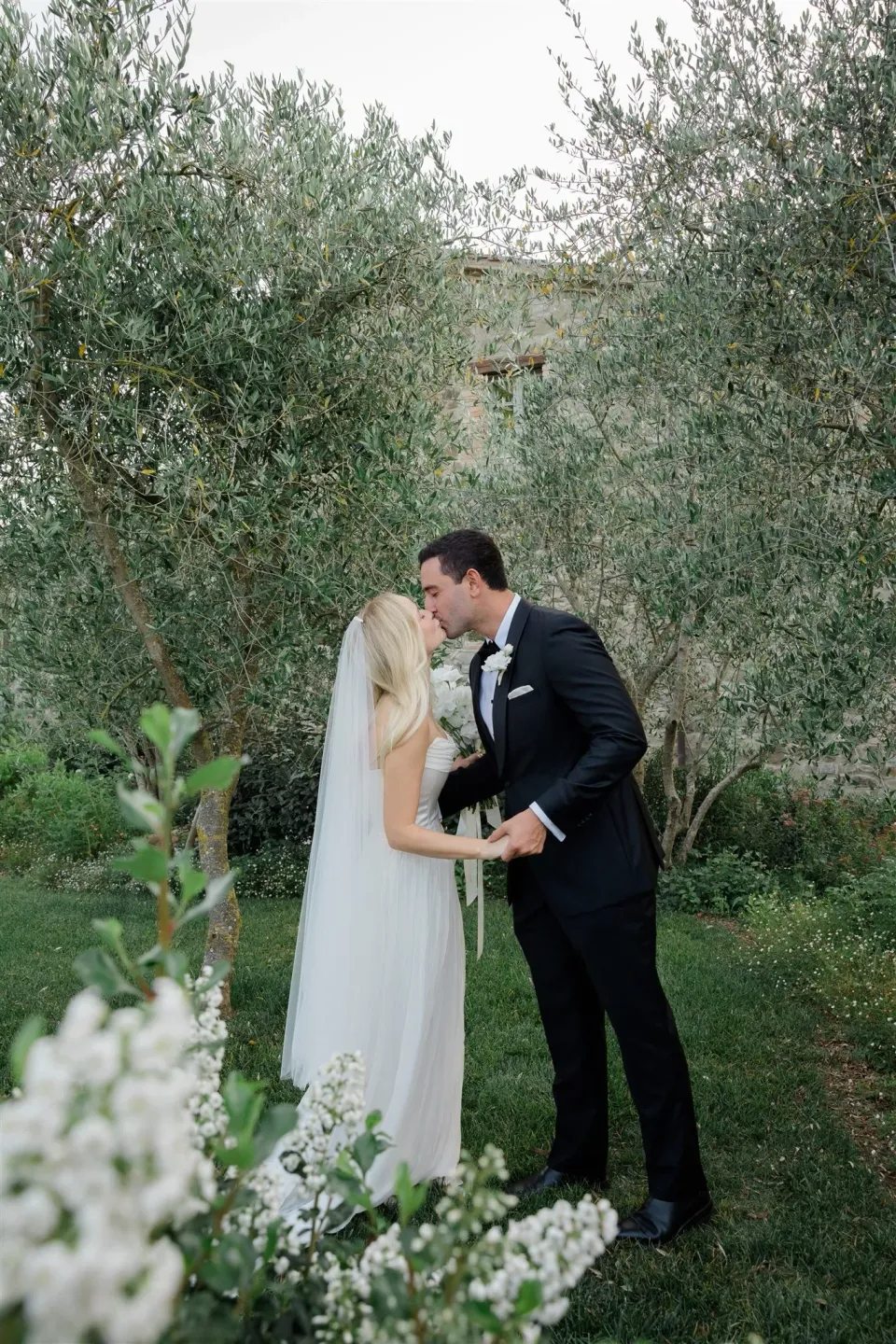 Shelby and Jack kissing among trees with white ceremony flowers by Le Francesche in the foreground, in Umbria, Italy, for an Elopement Collective elopement. Photo by James from House of Love.