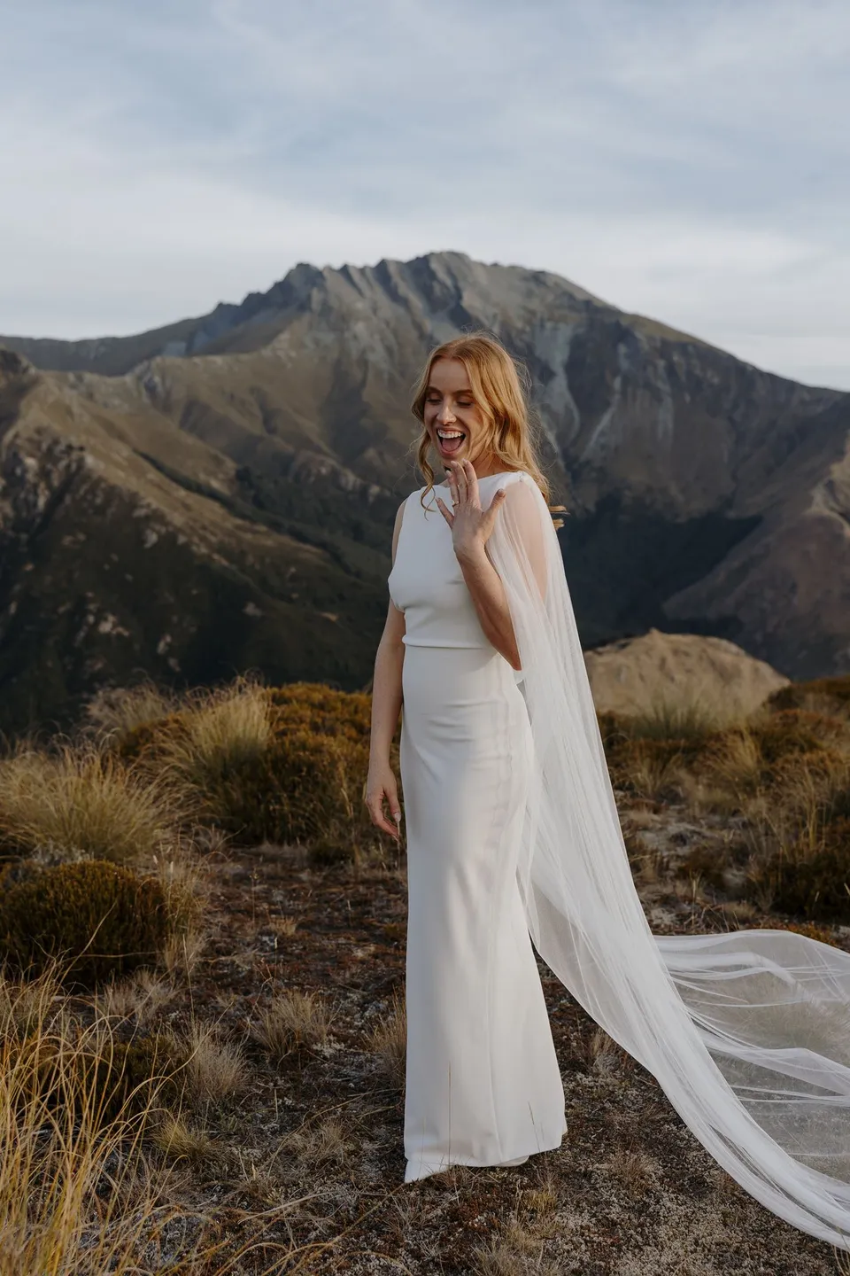 Neve smiling in her white wedding dress and flowing sheer cape, surrounded by golden tussock and mountain peaks, on Big Geordie above Queenstown, New Zealand, for an Elopement Collective Queenstown elopement. Photo by George Bowden.