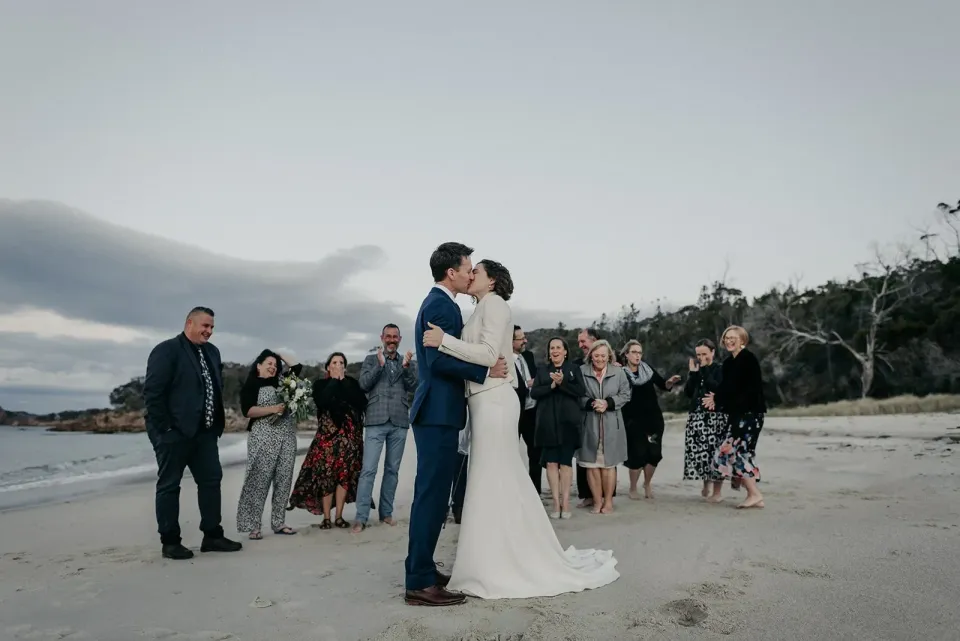A couple kiss on a beach in front of a small group of guests.