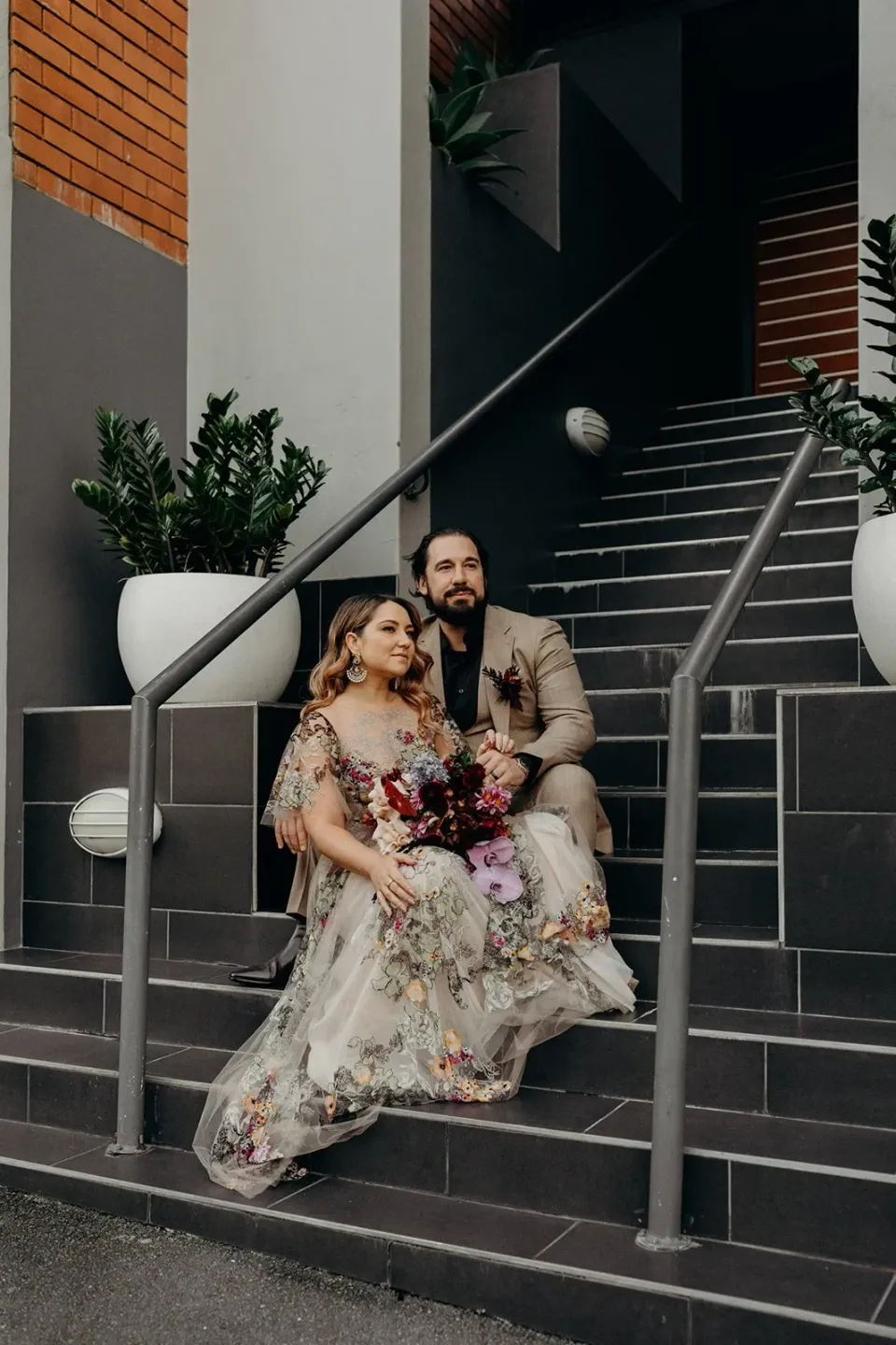 Victoria and Daniel sitting together on a flight of outdoor steps, her floral dress spread across the stair, in Fortitude Valley, Brisbane, for an Elopement Collective elopement. Photo by Tessa Shannon.