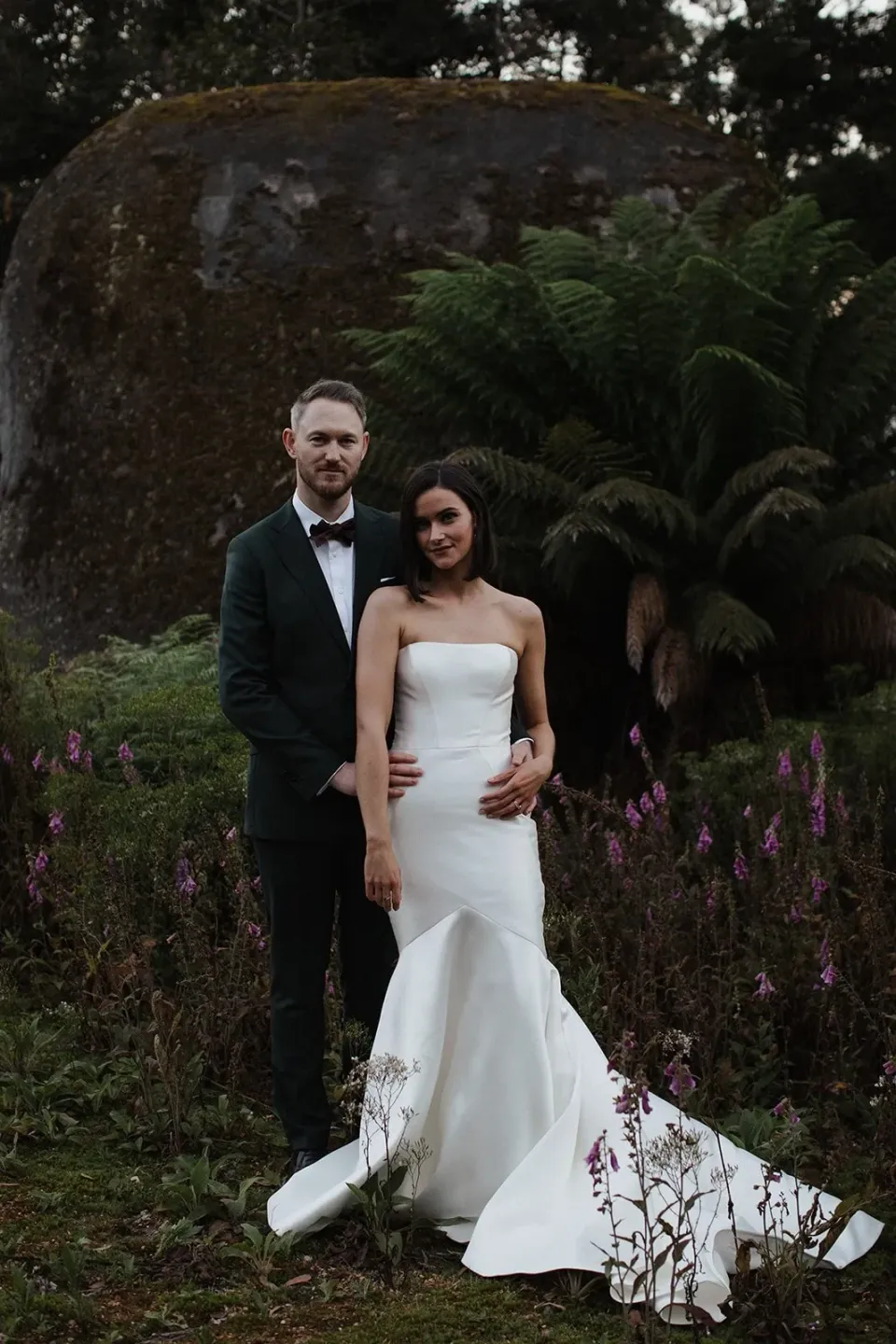 A couple stand together beside large boulders and green ferns.