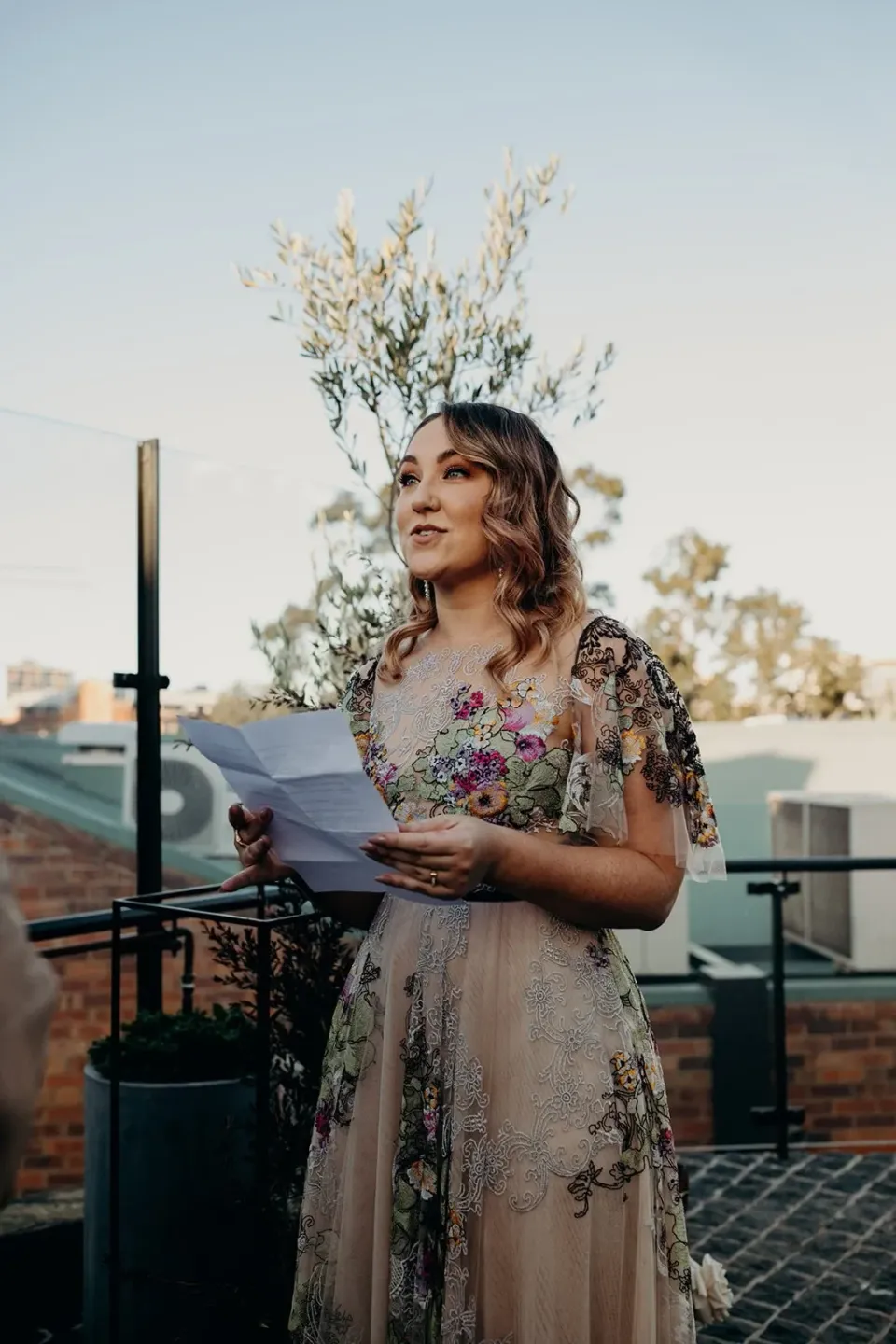 Victoria reading her vows from a sheet of paper on the rooftop terrace, at Agnes Restaurant in Fortitude Valley, Brisbane, for an Elopement Collective elopement. Photo by Tessa Shannon.