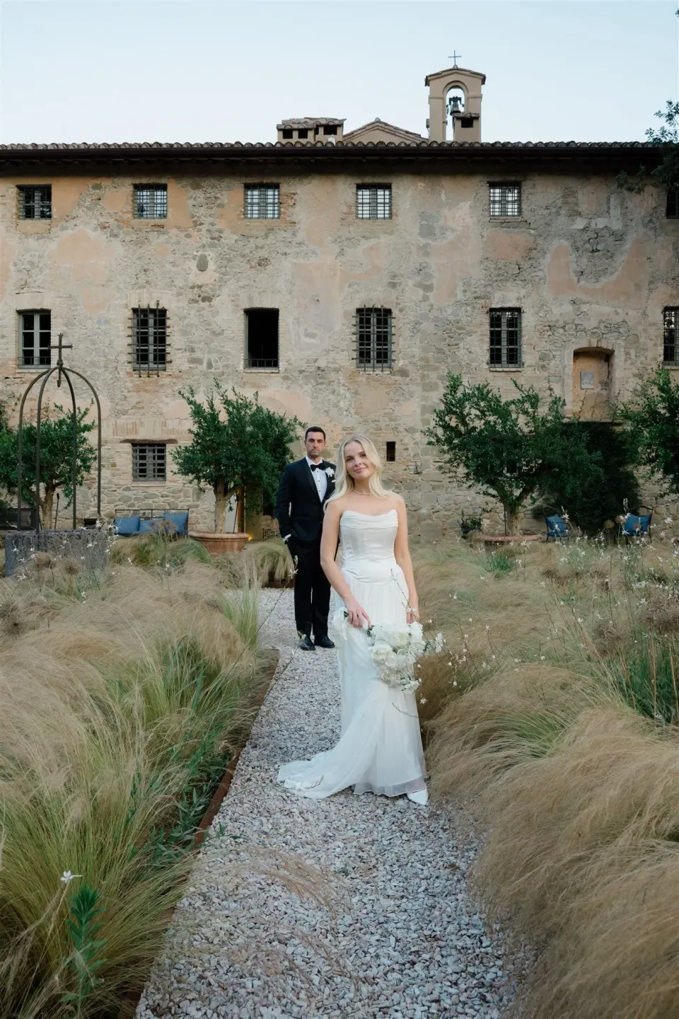 Shelby leading Jack along a gravel path through ornamental grasses, in Umbria, Italy, for an Elopement Collective elopement. Photo by James from House of Love.
