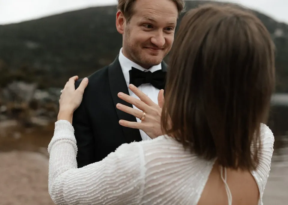 Josh smiling at April as she lifts her hands towards his shoulders, showing her wedding ring, at Dove Lake, Cradle Mountain, Tasmania, for an Elopement Collective elopement. Photo by Bulb Creative.