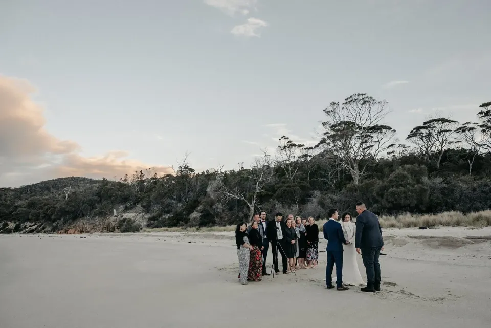 A small group gathers with a couple on a broad sandy beach at dusk.