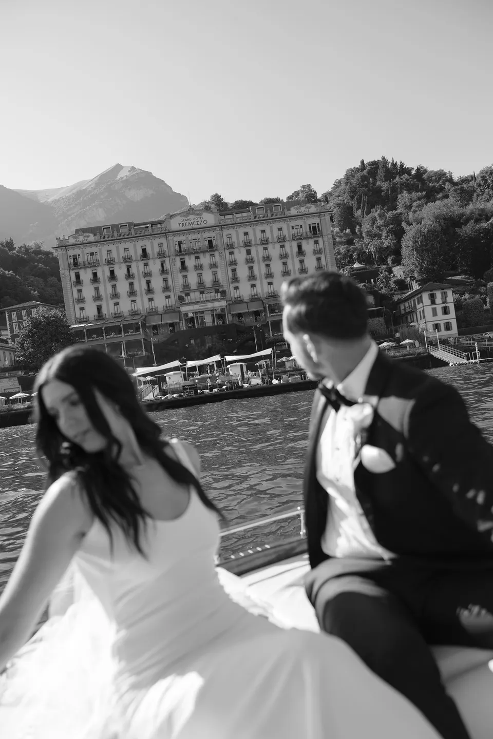 Black and white photograph of Lyndal and Lachlan smiling together on a boat, with a lakeside building and mountains behind them, at Lake Como in Lombardy, Italy, for an Elopement Collective elopement. Photograph by George Bowden.