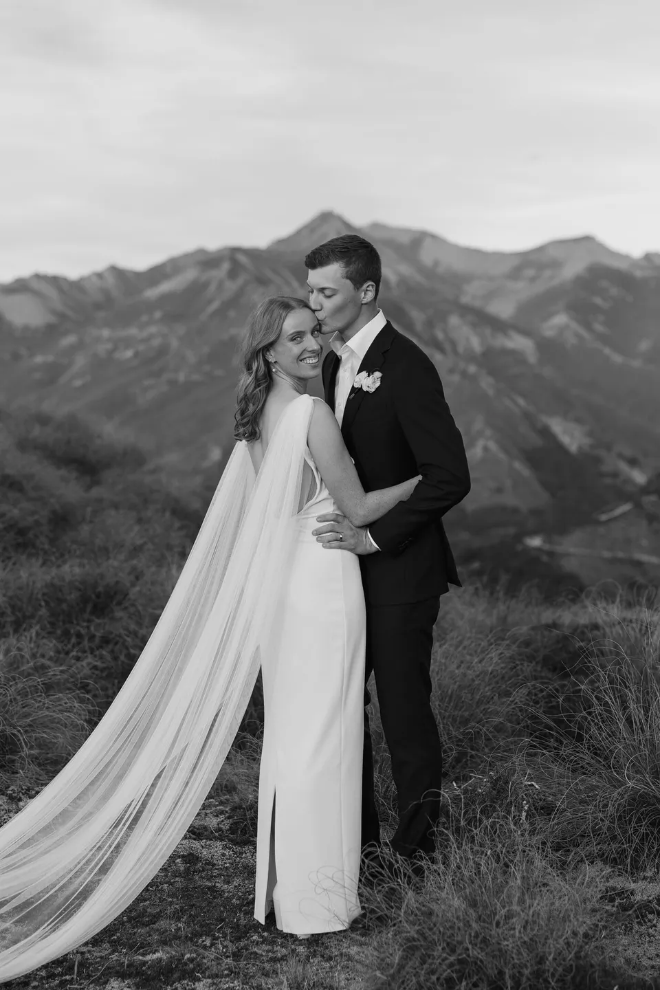 A black and white portrait of Neve and Hamish embracing, with her long cape stretching across the mountain grass, on Big Geordie above Queenstown, New Zealand, for an Elopement Collective Queenstown elopement. Photo by George Bowden.