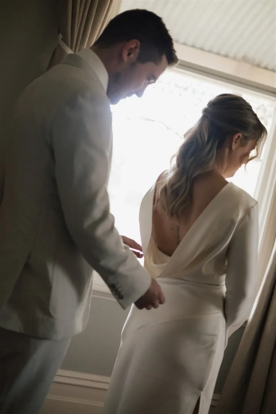 Cameron helping Maddy with the back of her white dress beside a window, at The Mayor’s Residence in Daylesford, Victoria, for an Elopement Collective elopement. Photo by Bulb Creative.