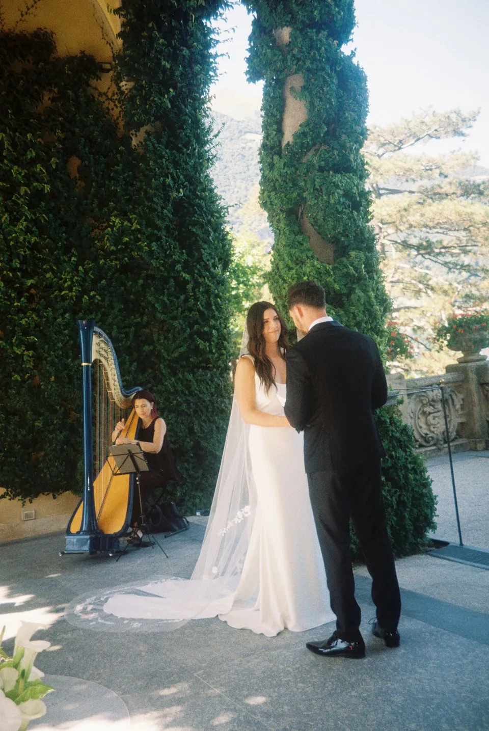 Lyndal facing Lachlan during their ceremony beneath Villa del Balbianello’s ivy loggia, with a harpist seated nearby, at Lake Como in Lombardy, Italy, for an Elopement Collective elopement. Photograph by George Bowden.