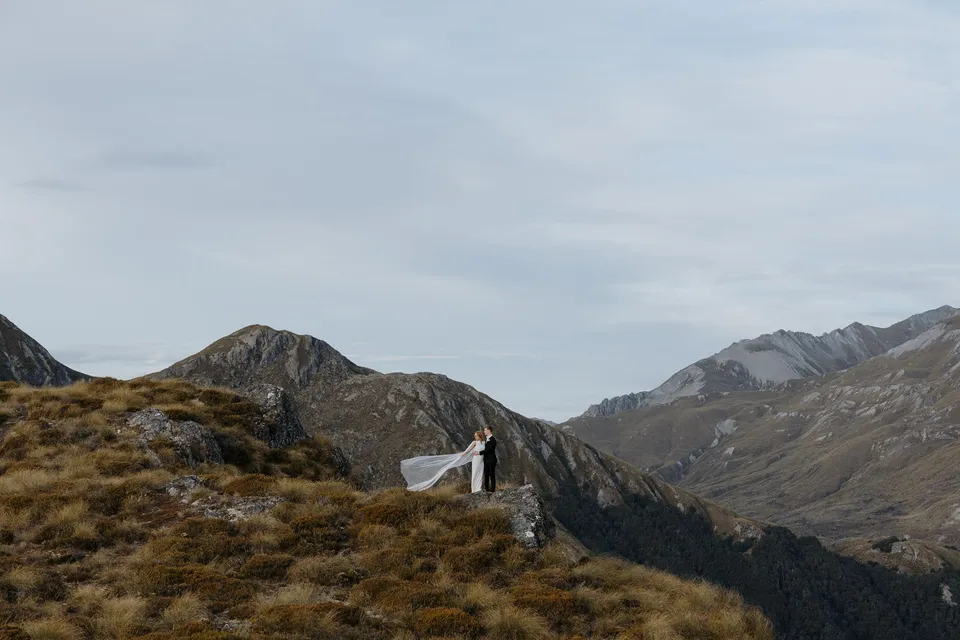 A wide view of Neve and Hamish standing on a tussock-covered ridge beneath an open sky and jagged mountain peaks, on Big Geordie above Queenstown, New Zealand, for an Elopement Collective Queenstown elopement. Photo by George Bowden.