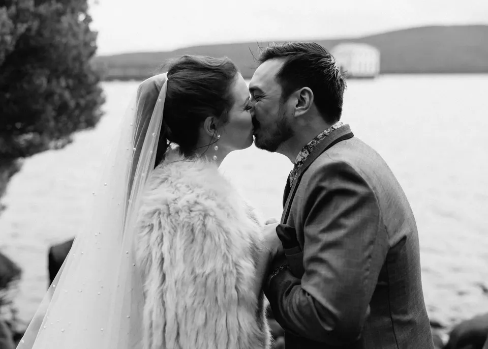 Black-and-white photograph of Jono kissing Sarah’s forehead by the lakeshore, at Pumphouse Point, Lake St Clair, Tasmania, for an Elopement Collective elopement. Photo by Bulb Creative.