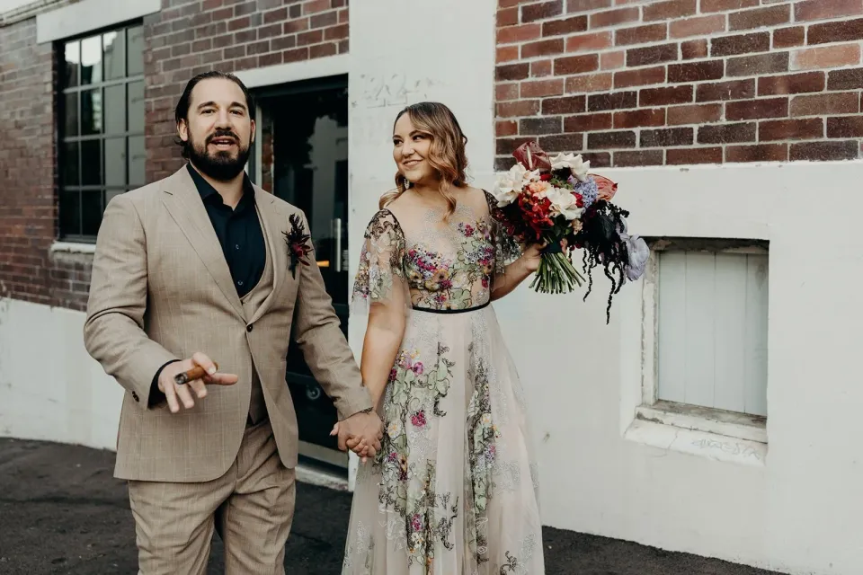Victoria and Daniel standing outside a brick building, with her colourful bouquet by Mrs Gibbons Flowers, in Fortitude Valley, Brisbane, for an Elopement Collective elopement. Photo by Tessa Shannon.