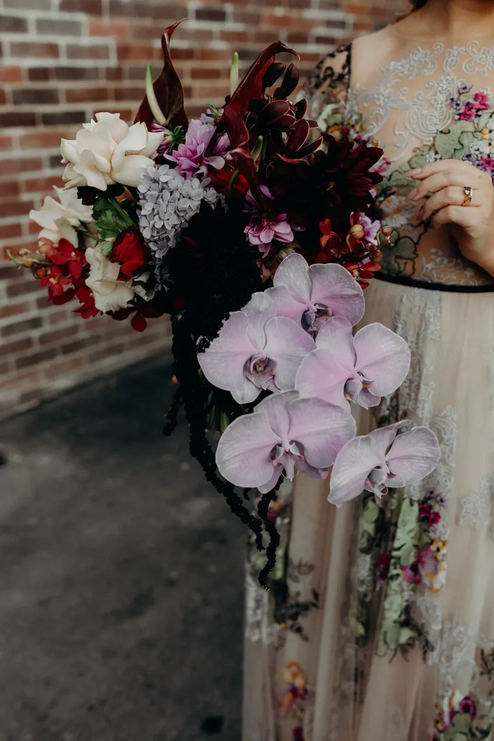 Close-up of Victoria holding her cascading bouquet by Mrs Gibbons Flowers against her floral dress, in Fortitude Valley, Brisbane, for an Elopement Collective elopement. Photo by Tessa Shannon.