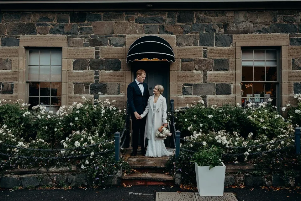 A couple stand in the doorway of a stone house surrounded by white flowering plants.