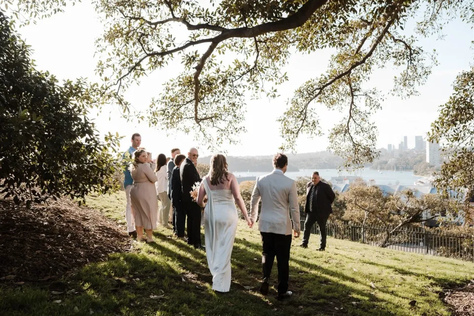 Alyce and Andrew approaching their guests beneath a spreading tree above Sydney Harbour, at Observatory Hill in Sydney, New South Wales, for an Elopement Collective elopement. Photo by Jason Corroto.