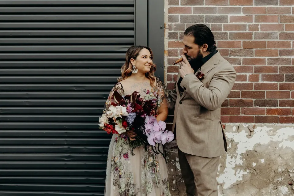 Victoria smiling at Daniel as he smokes a cigar beside her bouquet by Mrs Gibbons Flowers, in Fortitude Valley, Brisbane, for an Elopement Collective elopement. Photo by Tessa Shannon.