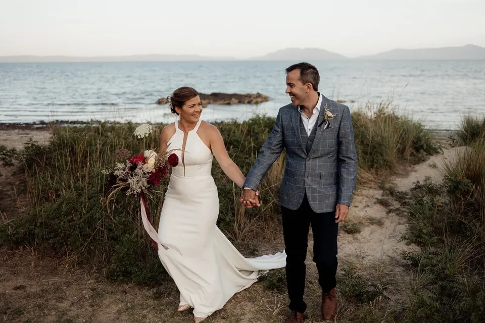 A couple walk hand in hand through coastal grass above the beach.