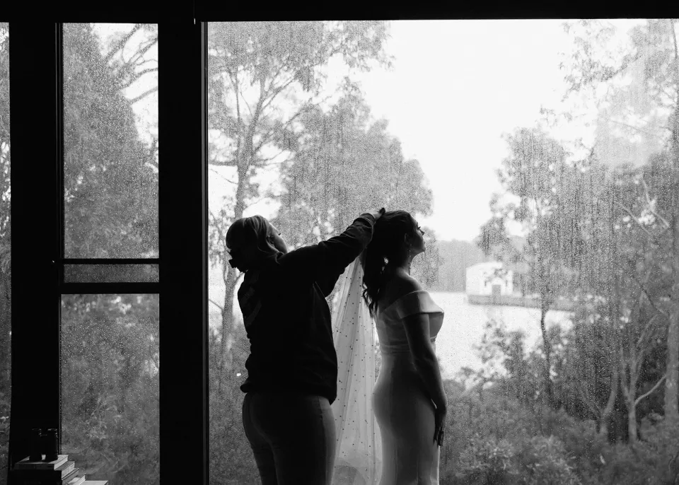 Black-and-white silhouette of a woman adjusting Sarah’s hair beside the window, at Pumphouse Point, Lake St Clair, Tasmania, for an Elopement Collective elopement. Photo by Bulb Creative.