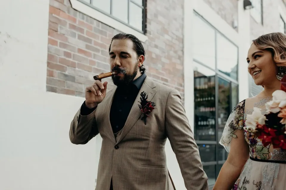 Daniel smoking a cigar beside Victoria and her bouquet by Mrs Gibbons Flowers, in Fortitude Valley, Brisbane, for an Elopement Collective elopement. Photo by Tessa Shannon.
