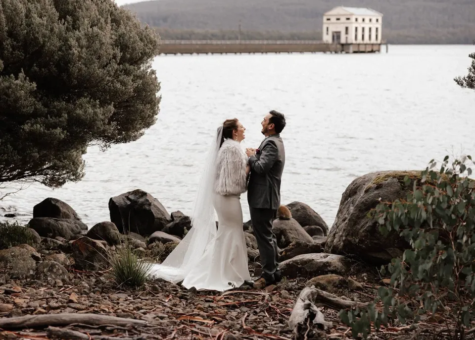 Sarah and Jono laugh together on the rocky shore of Lake St Clair, with Pumphouse Point across the water.
