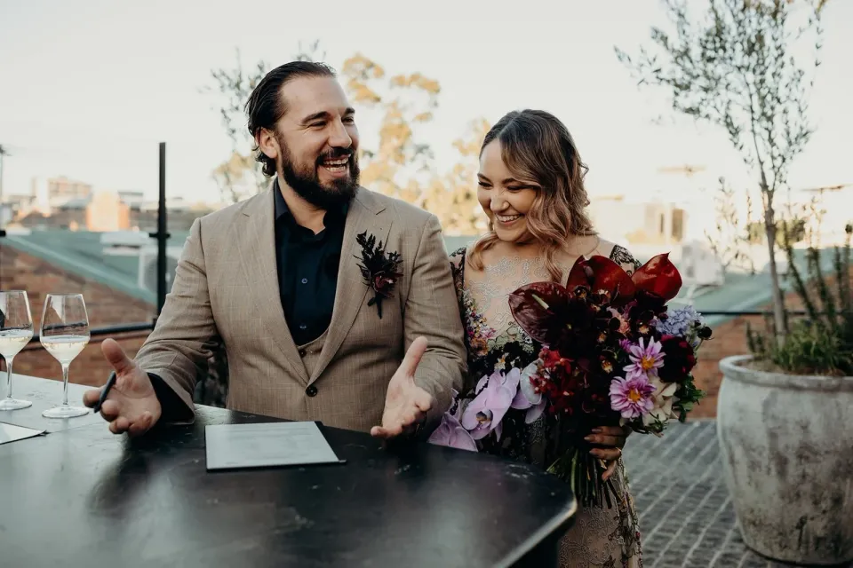 Victoria and Daniel laughing beside their signing paperwork and her bouquet by Mrs Gibbons Flowers, at Agnes Restaurant in Fortitude Valley, Brisbane, for an Elopement Collective elopement. Photo by Tessa Shannon.