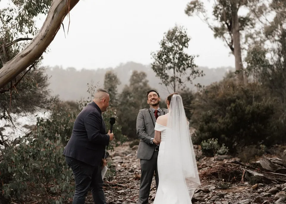 A couple laugh with their celebrant during a lakeside ceremony beneath a gum tree.