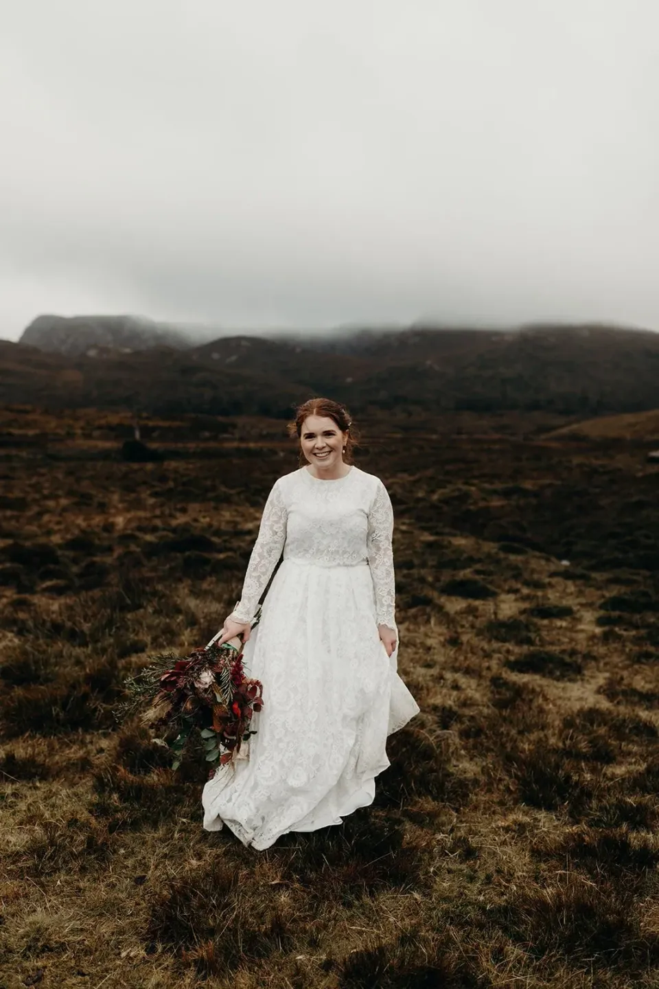 A bride walks through low alpine vegetation beneath misty hills.
