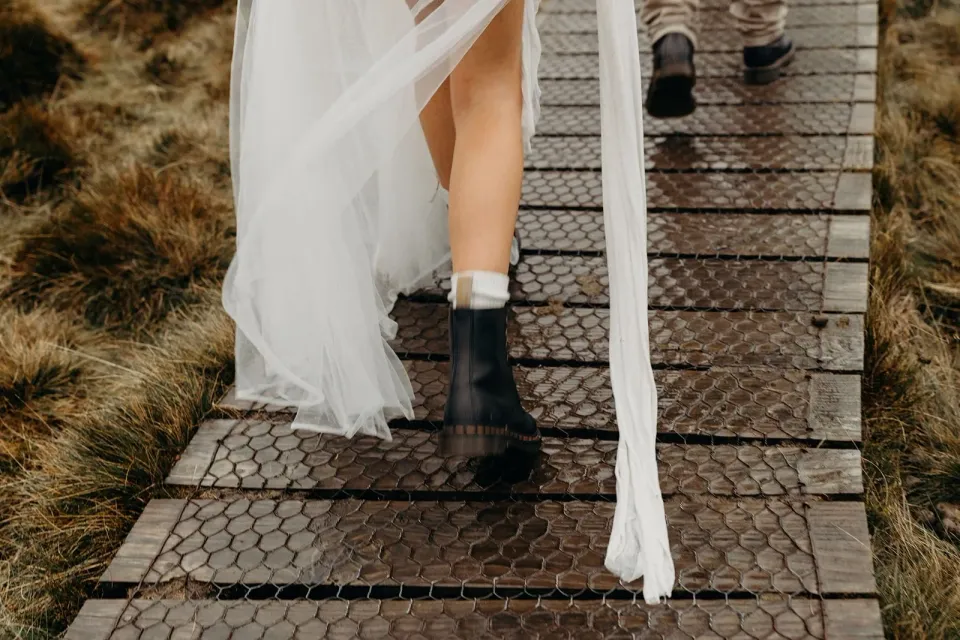 A bride's boots and flowing train on a timber boardwalk through alpine grass.