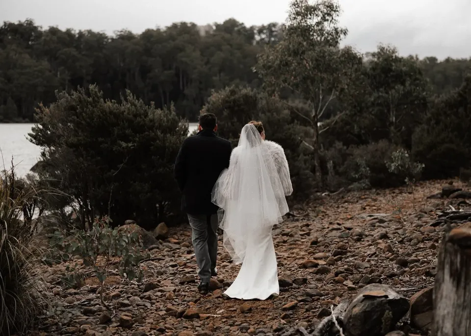 A couple walk along a leaf-covered lakeside path beneath trees.
