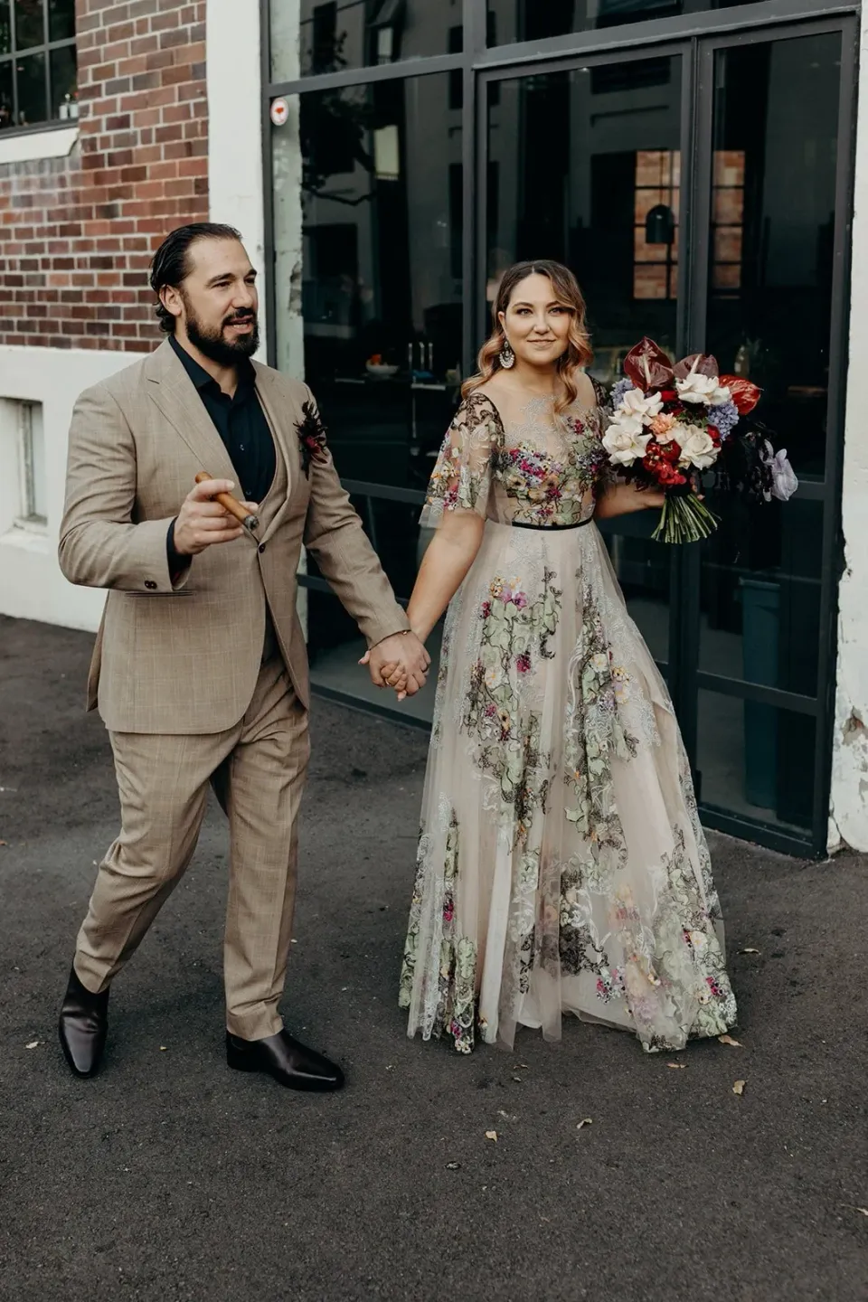 Full-length portrait of Victoria and Daniel holding hands outside a dark doorway, with her bouquet by Mrs Gibbons Flowers, in Fortitude Valley, Brisbane, for an Elopement Collective elopement. Photo by Tessa Shannon.