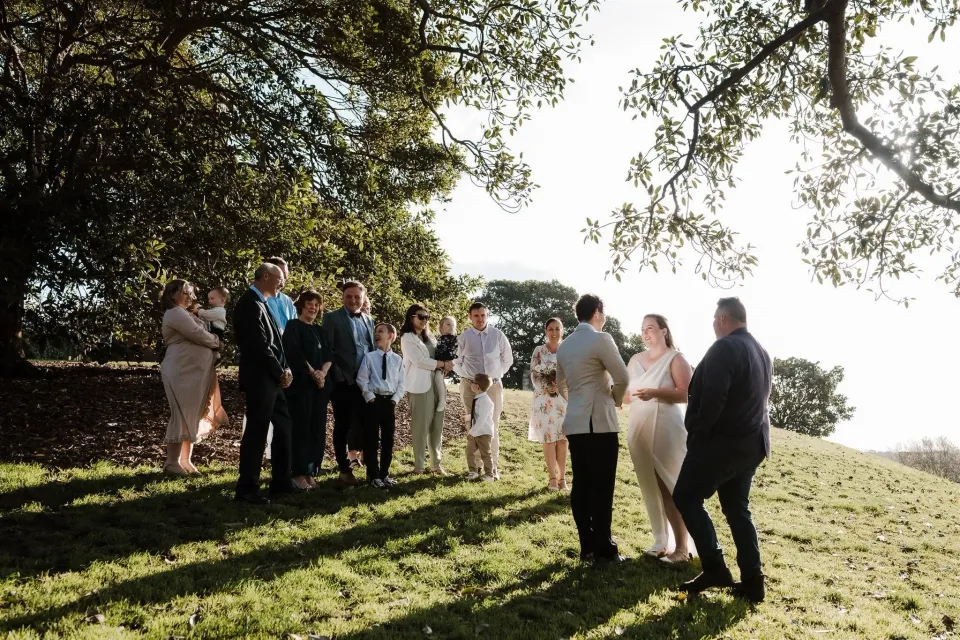 Alyce and Andrew exchanging vows with celebrant Josh Withers and guests gathered beneath a large tree, at Observatory Hill in Sydney, New South Wales, for an Elopement Collective elopement. Photo by Jason Corroto.