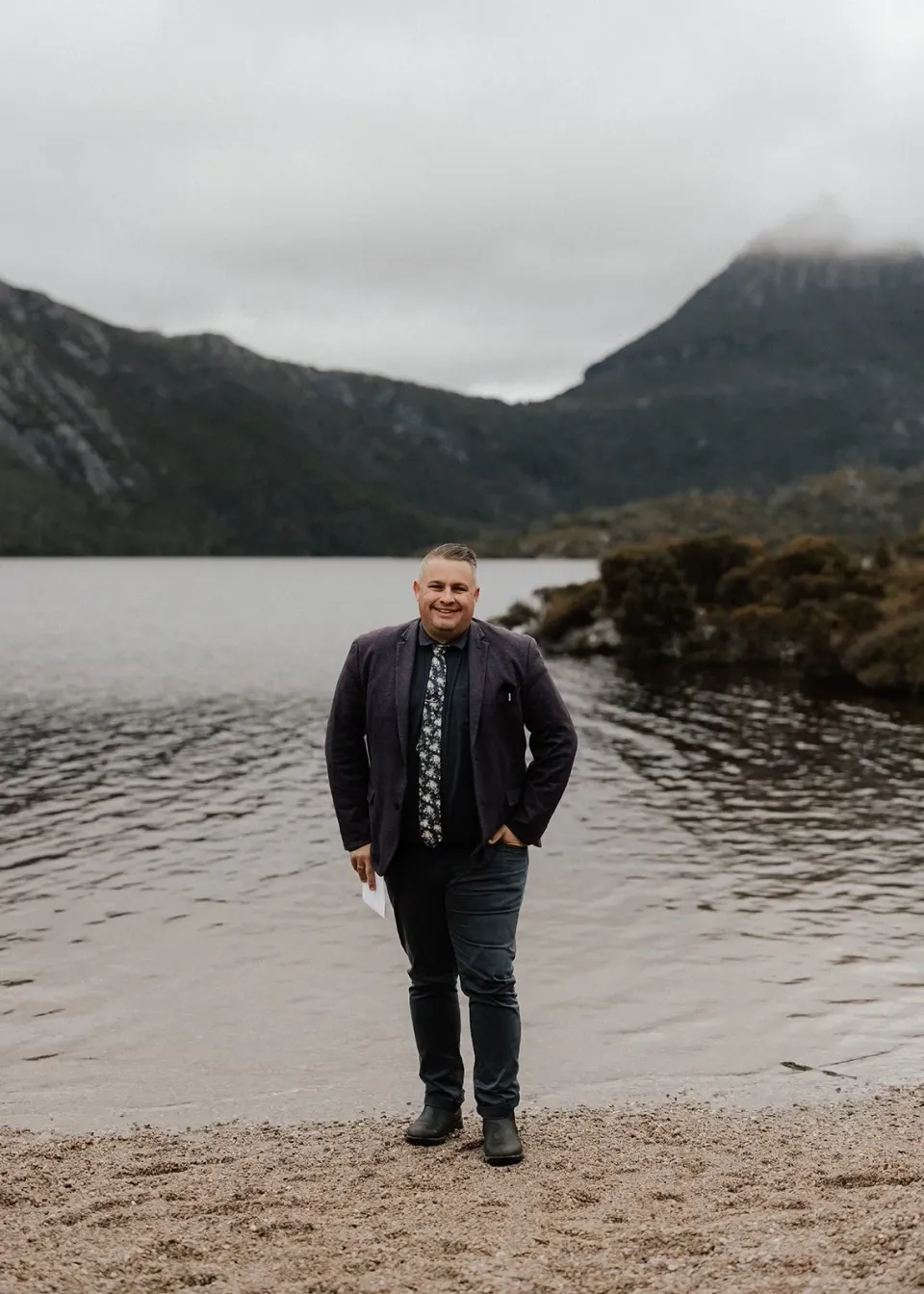 Celebrant Josh Withers standing on the lakeshore beneath misty mountain peaks, at Dove Lake, Cradle Mountain, Tasmania, for an Elopement Collective elopement. Photo by Bulb Creative.