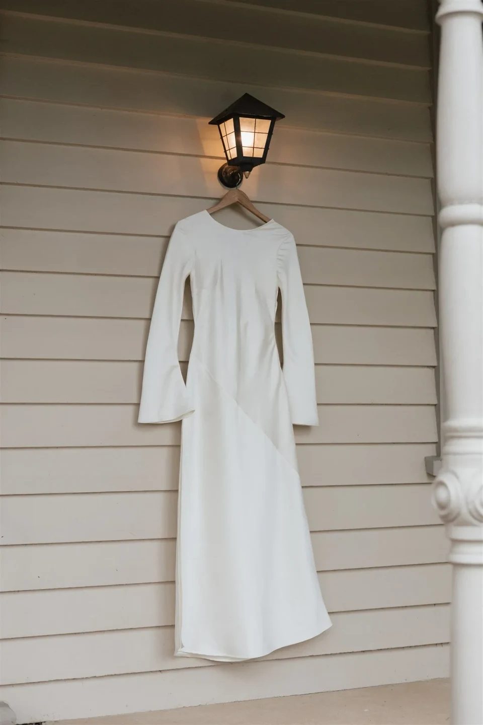 Maddy’s white Olivia Rose the Label dress hanging on a weatherboard veranda beneath a lantern, at The Mayor’s Residence in Daylesford, Victoria, for an Elopement Collective elopement. Photo by Bulb Creative.
