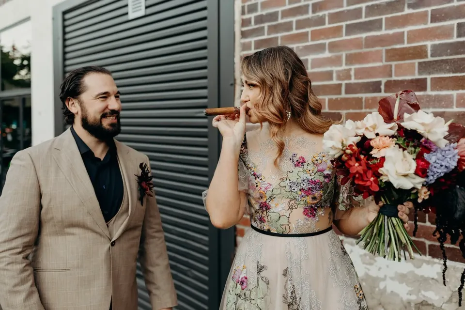 Victoria smoking a cigar while Daniel smiles beside her and her bouquet by Mrs Gibbons Flowers, in Fortitude Valley, Brisbane, for an Elopement Collective elopement. Photo by Tessa Shannon.