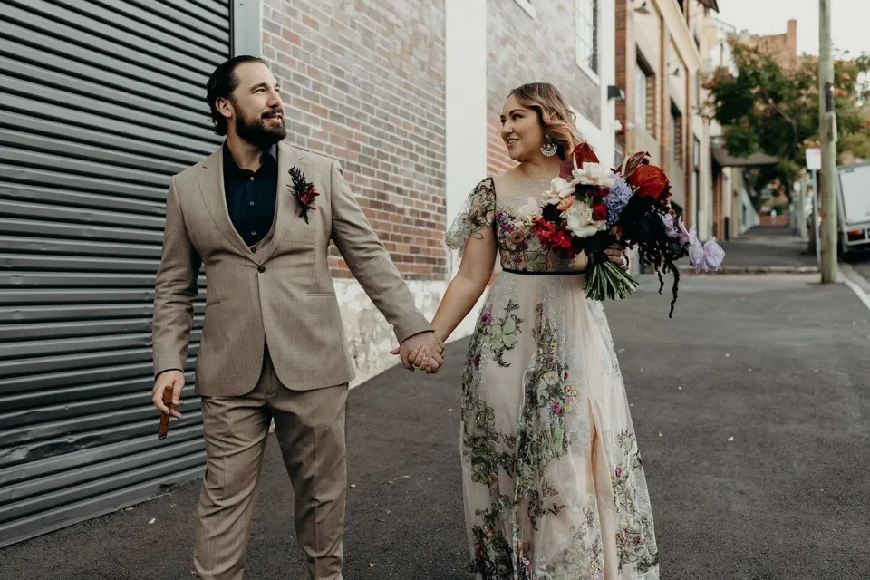 Victoria and Daniel holding hands on a city footpath beside a brick building, with her bouquet by Mrs Gibbons Flowers, in Fortitude Valley, Brisbane, for an Elopement Collective elopement. Photo by Tessa Shannon.