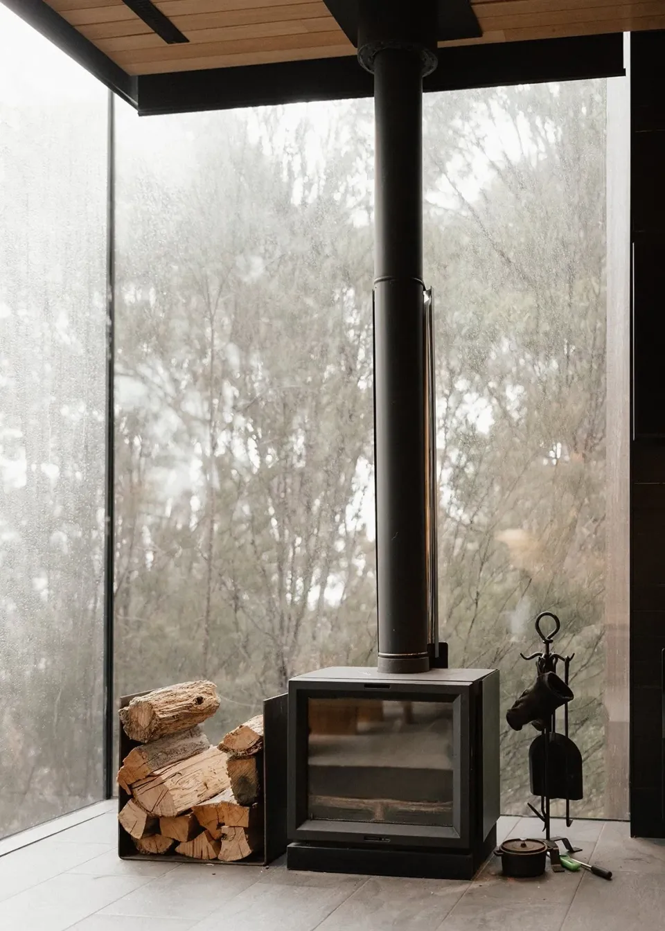 A wood-burning stove and stacked logs beside floor-to-ceiling windows looking into the trees, at Pumphouse Point, Lake St Clair, Tasmania, for an Elopement Collective elopement. Photo by Bulb Creative.