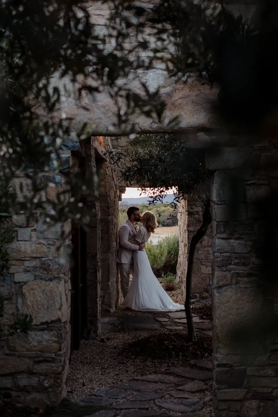 A couple kiss in a doorway framed by stone walls and leafy branches.